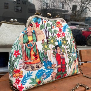 Può includere: Una pochette bianca con una chiusura a scatto dorata e una tracolla a catena. La pochette presenta una stampa floreale colorata con immagini di Frida Kahlo.