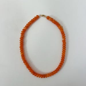 May include: A necklace composed of small, round, orange beads. The beads are strung together, forming a complete circle. The clasp is gold-colored. The necklace is displayed against a white background.
