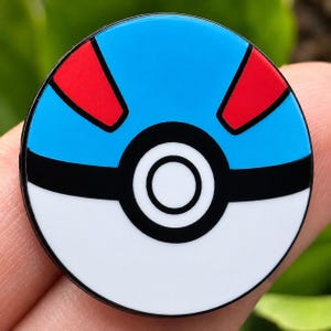 Puede incluir: Un pin esmaltado circular con un diseño estilizado de una Super Ball. La mitad superior es azul claro con dos acentos triangulares rojos, mientras que la mitad inferior es blanca. Una gruesa banda negra horizontal cruza el centro con un detalle de botón circular central en blanco y negro.