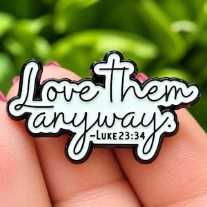 Puede incluir: Un pin de esmalte blanco con bordes y letras de metal negro que dice Love Them anyway -LUKE 23:34. El pin presenta una fuente cursiva para el texto principal y una fuente sans-serif limpia para la referencia bíblica sobre un fondo blanco.