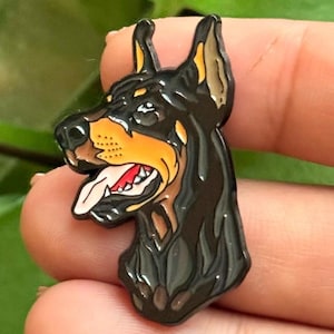 May include: A black and tan Doberman dog enamel pin featuring a profile view of the canine with its mouth open, tongue out, and ears pointed upward. The design uses bold black outlines, a tan muzzle, and a white tongue with red accents.