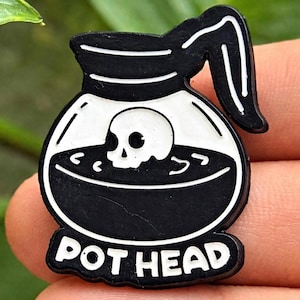 May include: A black and white enamel pin shaped like a coffee pot containing a skull submerged in dark liquid. The design features a handle on the right side and the text POT HEAD printed in bold white capital letters along the bottom edge.