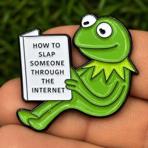 May include: A bright green frog enamel pin featuring a white book with black text that reads HOW TO SLAP SOMEONE THROUGH THE INTERNET. The frog has large white eyes with black pupils and a pointed collar, rendered in a bold, cartoon-style design.