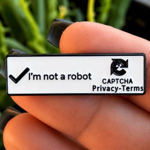 May include: A rectangular enamel pin with a white background and black border. The pin features a black checkmark icon, the text "I'm not a robot", a circular refresh arrow icon, and the text "CAPTCHA Privacy-Terms" printed in black lettering.