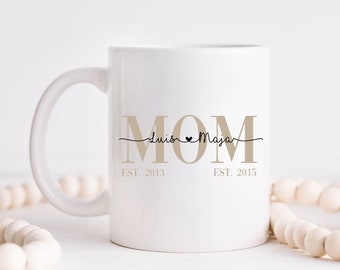 Könnte beinhalten: Weiße Keramiktasse mit dem Wort "MOM" in brauner Schrift. Der Text "Luis ♥ Maya" ist in schwarzer Schrift unter dem Wort "MOM" geschrieben. Die Texte "EST. 2013" und "EST. 2015" sind unter der Schrift geschrieben.