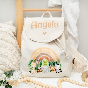 Può includere: Uno zaino in tela beige con un design a arcobaleno e stampa di animali. Lo zaino ha una chiusura a coulisse e un piccolo cuore sopra il nome "Angelo" stampato sul davanti.