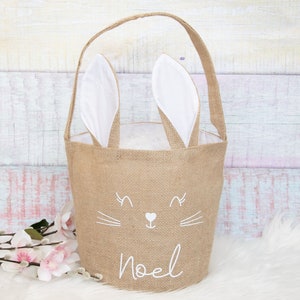Può includere: Un cestino di Pasqua in juta marrone con orecchie di coniglio e un viso di coniglio ricamato in bianco. Il cestino ha la scritta "Noel" sul davanti.