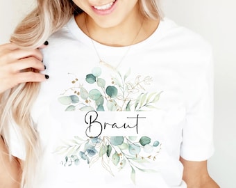 May include: White t-shirt with a green and gold floral design and the word "Braut" in black script.
