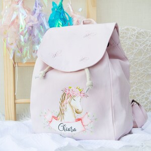 Può includere: Uno zaino in tela rosa con un designo di unicorno e il nome "Chiara" stampato su uno striscione. L'unicorno indossa una corona di fiori ed è circondato da verde.