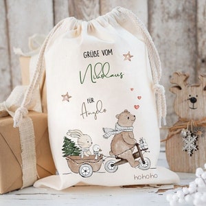 May include: A cream-colored drawstring gift bag with a whimsical illustration of a bear riding a tricycle and pulling a cart with a bunny and a duck. The bag has text in green and black, and is decorated with stars and hearts.