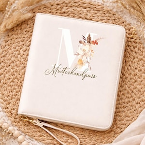 Könnte beinhalten: Cremefarbener Dokumentenhalter mit Reißverschluss, dem Buchstaben "M" und floralem Design. Der Text "Mutterkindpass" ist auf der Vorderseite aufgedruckt. Der Halter hat eine Handschlaufe und einen goldfarbenen Reißverschluss.