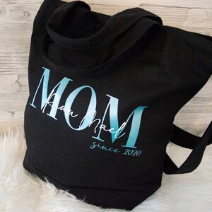 以下が含まれることがあります： 「MOM」と手書きの名前、その下に「since 2020」と書いてある、青と白のグラデーションデザインの黒いキャンバス地のトートバッグです。