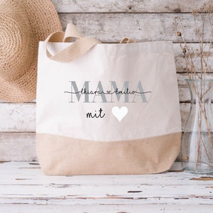 Pode incluir: Uma bolsa de lona bege com a palavra "MAMA" em letras cinzas e o texto "Chiara & Emilio" em escrita cursiva preta acima. A bolsa tamb&eacute;m tem a palavra "mit" em escrita cursiva preta e um cora&ccedil;&atilde;o branco.