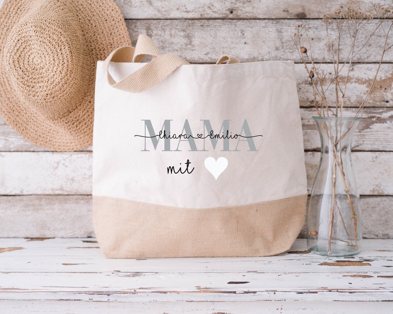 Pode incluir: Uma bolsa de lona bege com a palavra "MAMA" em letras cinzas e o texto "Chiara & Emilio" em escrita cursiva preta acima. A bolsa tamb&eacute;m tem a palavra "mit" em escrita cursiva preta e um cora&ccedil;&atilde;o branco.