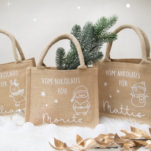 May include: Three burlap tote bags with rope handles. Each bag has a white illustration and text: a bunny, Santa Claus, and a snowman. The text reads "Vom Nikolaus f&uuml;r" followed by a name. A sprig of greenery is in one bag.