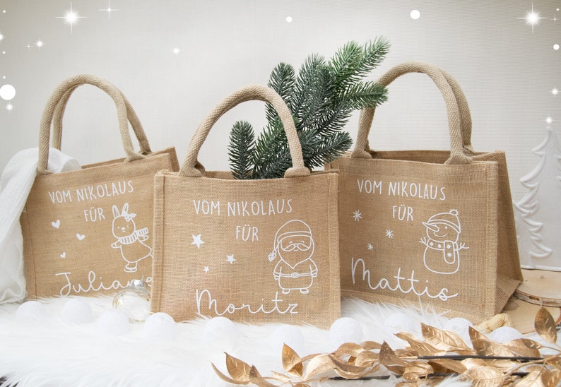 May include: Three burlap tote bags with rope handles. Each bag has a white illustration and text: a bunny, Santa Claus, and a snowman. The text reads "Vom Nikolaus f&uuml;r" followed by a name. A sprig of greenery is in one bag.