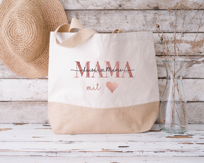 Pode incluir: Uma bolsa tote de lona bege com um design de glitter rosa que diz "MAMA" com os nomes "Luisa" e "Marie" escritos em it&aacute;lico acima da palavra "MAMA". A bolsa tamb&eacute;m tem um cora&ccedil;&atilde;o rosa e a palavra "mit" escrita abaixo da palavra "MAMA".