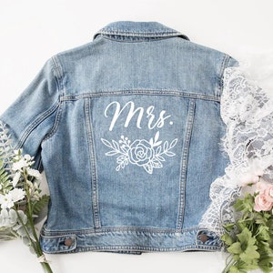 Peut inclure: Une veste en jean bleu clair avec un motif floral blanc et le mot "Mme" au dos.