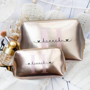 Op de afbeelding: Twee gouden toilettassen met een roze en witte monogram van de letter "H" en de naam "hannah" in zwarte schrift. De tassen liggen op een wit oppervlak.