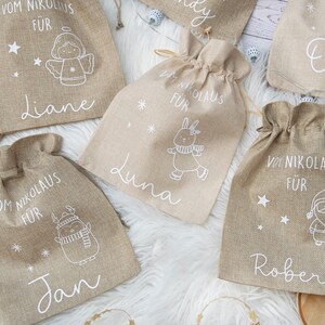 May include: Several burlap gift bags with drawstring closures are displayed. Each bag features a white illustration of a holiday character and a name. The text "Vom Nikolaus F&uuml;r" is printed above each name. The bags are tan.