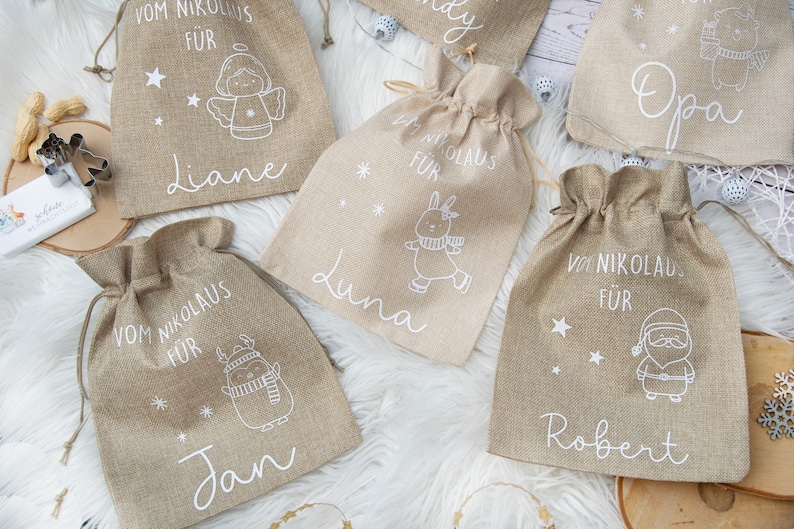May include: Several burlap gift bags with drawstring closures are displayed. Each bag features a white illustration of a holiday character and a name. The text "Vom Nikolaus F&uuml;r" is printed above each name. The bags are tan.