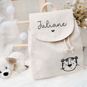 Può includere: Uno zaino in tela bianca con un design a forma di tigre nera e il nome "Juliane" scritto in corsivo nero con un piccolo cuore sopra.