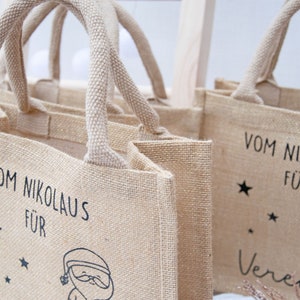 May include: Tan burlap tote bags with woven handles. Each bag features black text that reads "Vom Nikolaus F&uuml;r" and a Santa Claus illustration. One bag is personalized with a name. Stars are also included in the design.