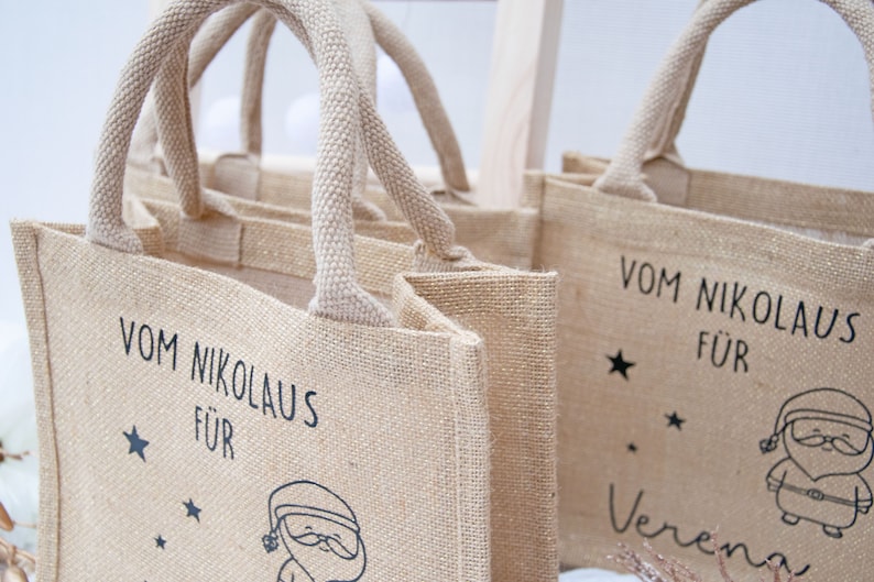 May include: Tan burlap tote bags with woven handles. Each bag features black text that reads "Vom Nikolaus F&uuml;r" and a Santa Claus illustration. One bag is personalized with a name. Stars are also included in the design.