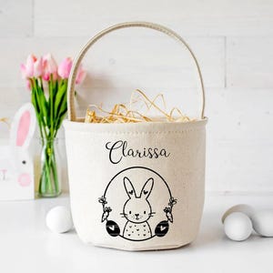 May include: A white canvas Easter basket with a black bunny and flower design. The basket has a handle and is filled with straw. The name "Clarissa" is printed on the front of the basket.