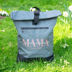 May include: Gray and black roll-top backpack with a zippered pocket and a pink glitter design that says "MAMA Martina & Nicola since 2008".