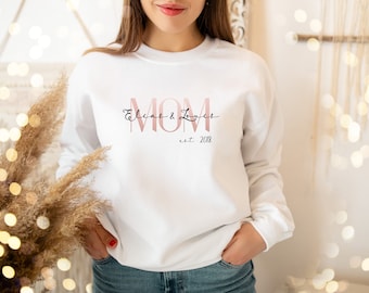Pode incluir: Camisola branca com a palavra "MOM" em rosa, com os nomes "Elias & Loues" em itálico acima e "est. 2018" abaixo. A camisola é de gola redonda e manga comprida.