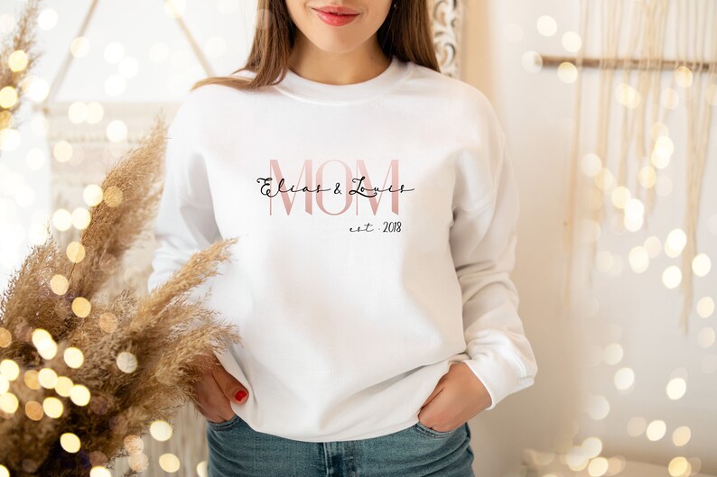May include: White sweatshirt with the word "MOM" in a pink font, with the names "Elias & Loues" in a cursive font above and "est. 2018" below. The sweatshirt is a long-sleeved crew neck.