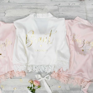 Könnte beinhalten: Drei Satin-Roben mit Spitzenbesatz. Die Roben sind rosa und weiß und haben goldene Beschriftungen. Die weiße Robe sagt "Bride" und die rosa Roben sagen "Bridesmaid" und "Maid of Honor".