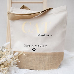 Puede incluir: Una bolsa de lona blanca con el texto "CAT mom" en letras doradas y "GIZMO & MARLEY" en letras negras con una huella de pata debajo. La parte inferior de la bolsa está hecha de arpillera.