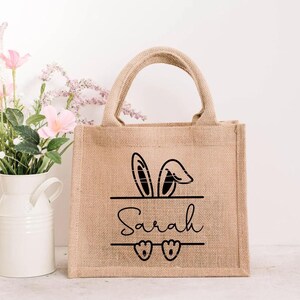 May include: A brown burlap tote bag with black bunny ears and the name "Sarah" printed on the front. The bag has two handles and is perfect for carrying small items.