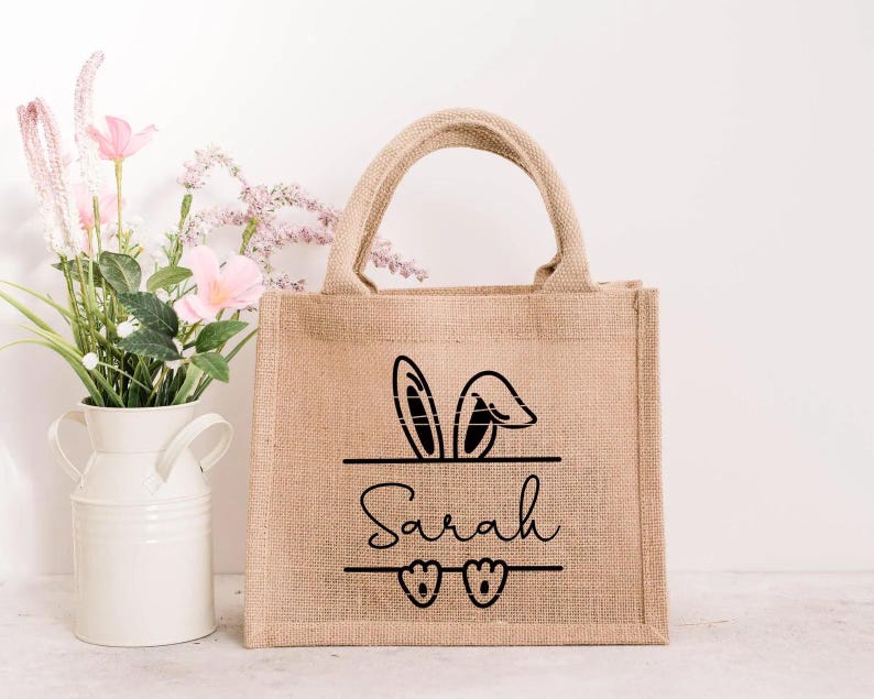 May include: A brown burlap tote bag with black bunny ears and the name "Sarah" printed on the front. The bag has two handles and is perfect for carrying small items.