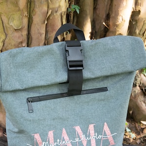 May include: Gray roll-top backpack with a black buckle and zipper. The backpack has the word "MAMA" with the names "Martina & Nicola" written underneath in pink glitter.
