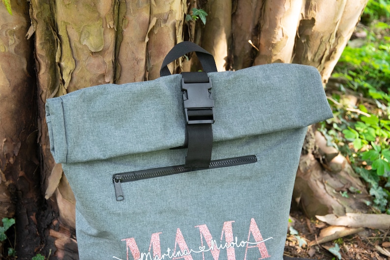May include: Gray roll-top backpack with a black buckle and zipper. The backpack has the word "MAMA" with the names "Martina & Nicola" written underneath in pink glitter.
