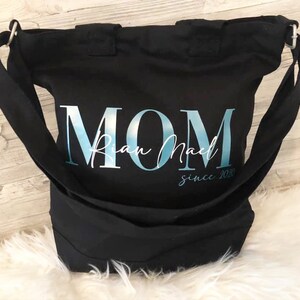 以下が含まれることがあります： 「MOM」と「Rian Mae」「since 2020」の文字が書かれた、青と白のデザインの黒いキャンバス地のトートバッグです。