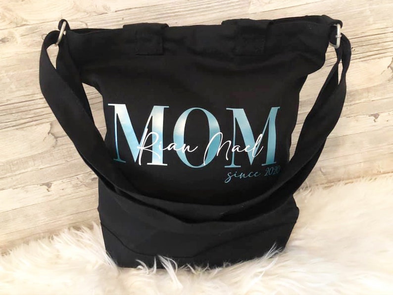 以下が含まれることがあります： 「MOM」と「Rian Mae」「since 2020」の文字が書かれた、青と白のデザインの黒いキャンバス地のトートバッグです。
