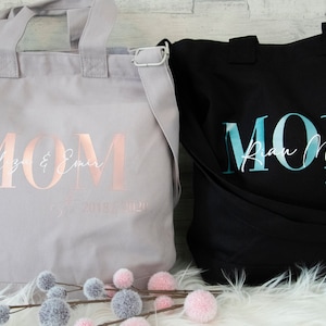 以下が含まれることがあります： 「MOM」とプリントされたキャンバス地のトートバッグ2つ。左側のバッグは薄いグレーで、ローズゴールドの文字で「Eliza & Emer since 2020」と書いてある。右側のバッグは黒で、ターコイズブルーの文字で「Ryan Noel since 2020」と書いてある。
