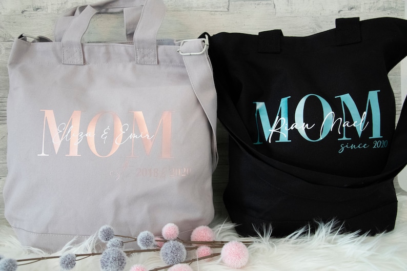 以下が含まれることがあります： 「MOM」とプリントされたキャンバス地のトートバッグ2つ。左側のバッグは薄いグレーで、ローズゴールドの文字で「Eliza & Emer since 2020」と書いてある。右側のバッグは黒で、ターコイズブルーの文字で「Ryan Noel since 2020」と書いてある。
