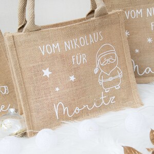 May include: Three burlap tote bags with white illustrations and text. One bag features a Santa Claus design, another a snowman, and the third a bunny. The text on the bags reads "Vom Nikolaus f&uuml;r" and individual names.