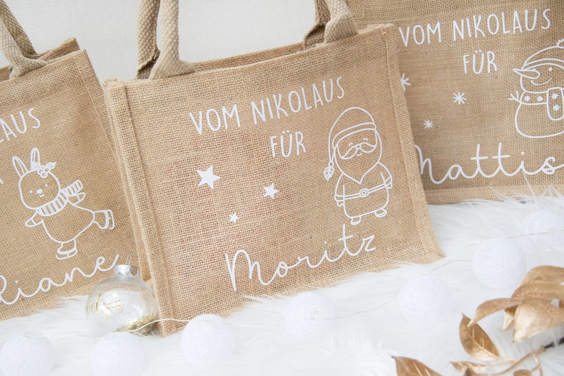 May include: Three burlap tote bags with white illustrations and text. One bag features a Santa Claus design, another a snowman, and the third a bunny. The text on the bags reads "Vom Nikolaus f&uuml;r" and individual names.