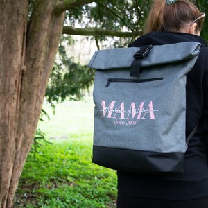 May include: Gray roll-top backpack with a black zipper and buckle. The backpack has a pink embroidered design that says "MAMA since 2008".