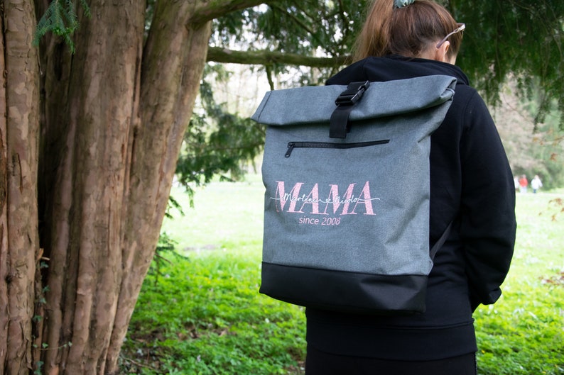 May include: Gray roll-top backpack with a black zipper and buckle. The backpack has a pink embroidered design that says "MAMA since 2008".