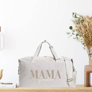 Puede incluir: Una bolsa de viaje de lona beige con una correa marr&oacute;n y el texto "MAMA Pauline" impreso en la parte delantera.