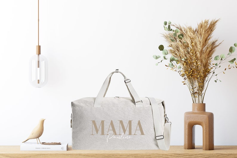 Puede incluir: Una bolsa de viaje de lona beige con una correa marr&oacute;n y el texto "MAMA Pauline" impreso en la parte delantera.