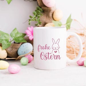 May include: White ceramic mug with a pink bunny silhouette and the text "Frohe Ostern" in pink script. The mug is surrounded by pastel Easter eggs and a wreath of greenery.