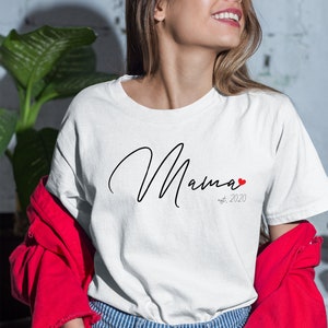 Op de afbeelding: Wit T-shirt met een zwarte, cursieve tekst die "Mama est. 2020" zegt met een klein rood hartje.